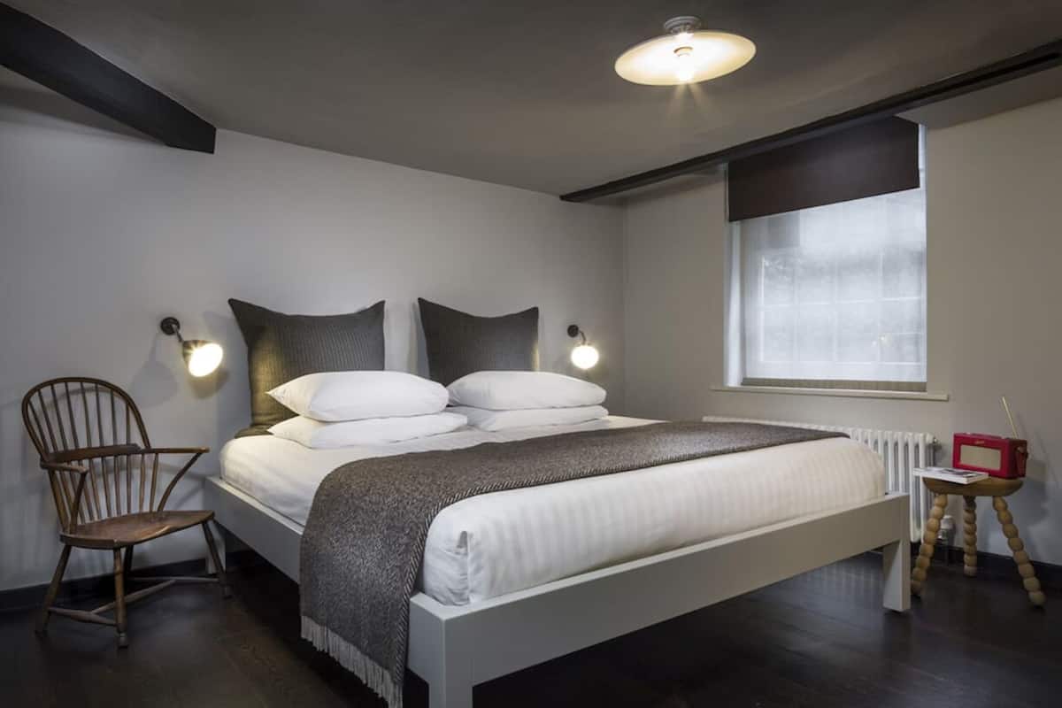 A modern bedroom with a large bed, a wooden chair, a bedside table with a radio, and a window with blinds.