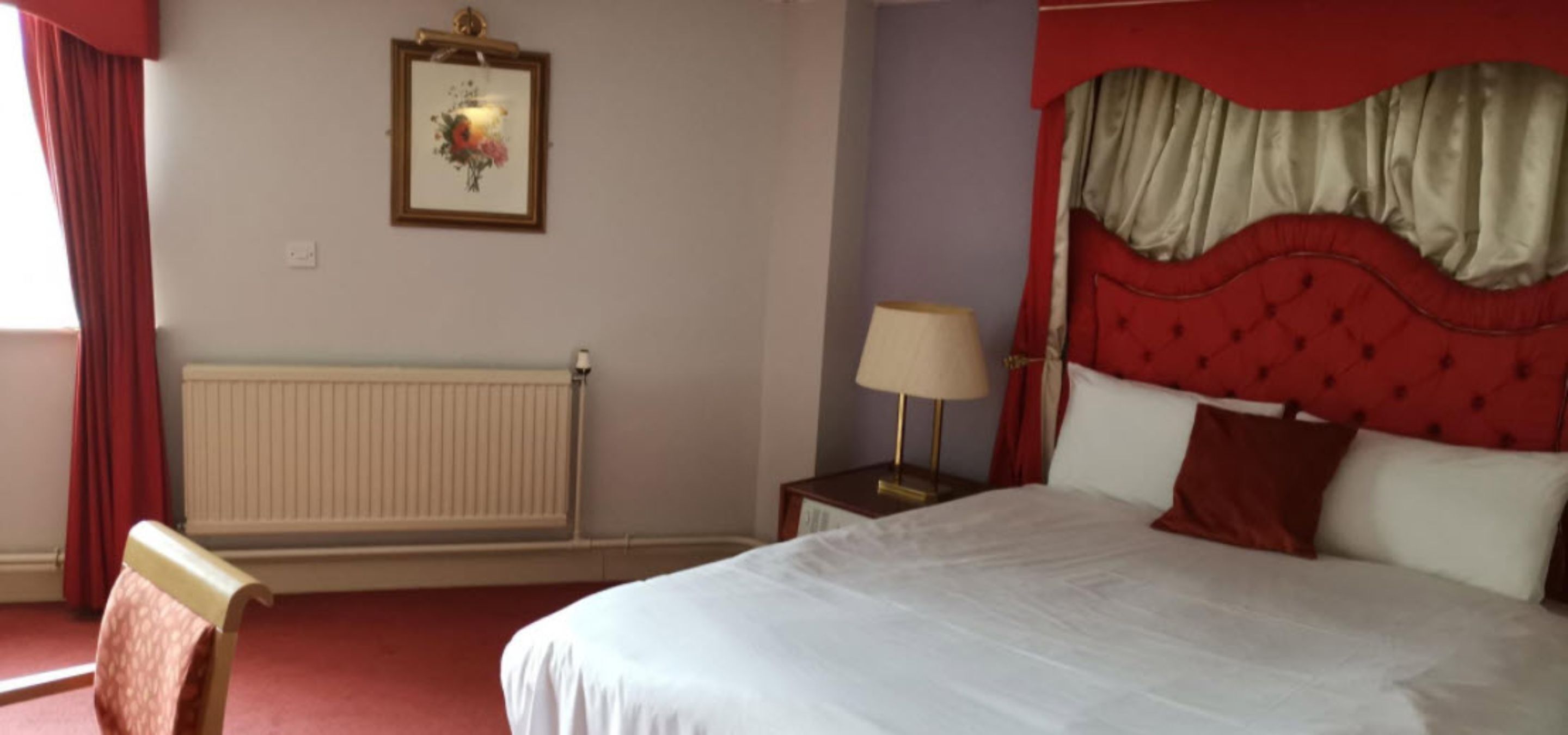 Una habitación de hotel con una cama, una mesita de noche con lámpara, un radiador y un cuadro en la pared.