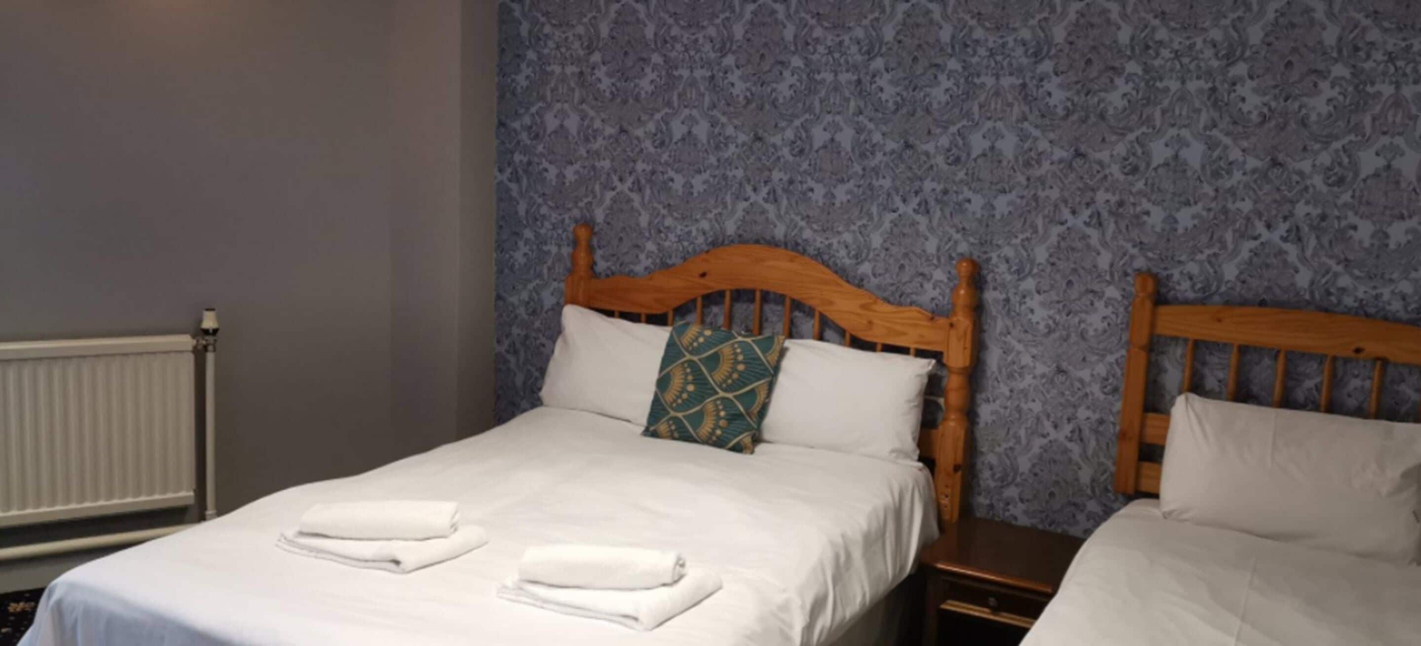 Cama de casal com lençóis brancos e duas toalhas dobradas, cabeceira de madeira e papel de parede estampado.