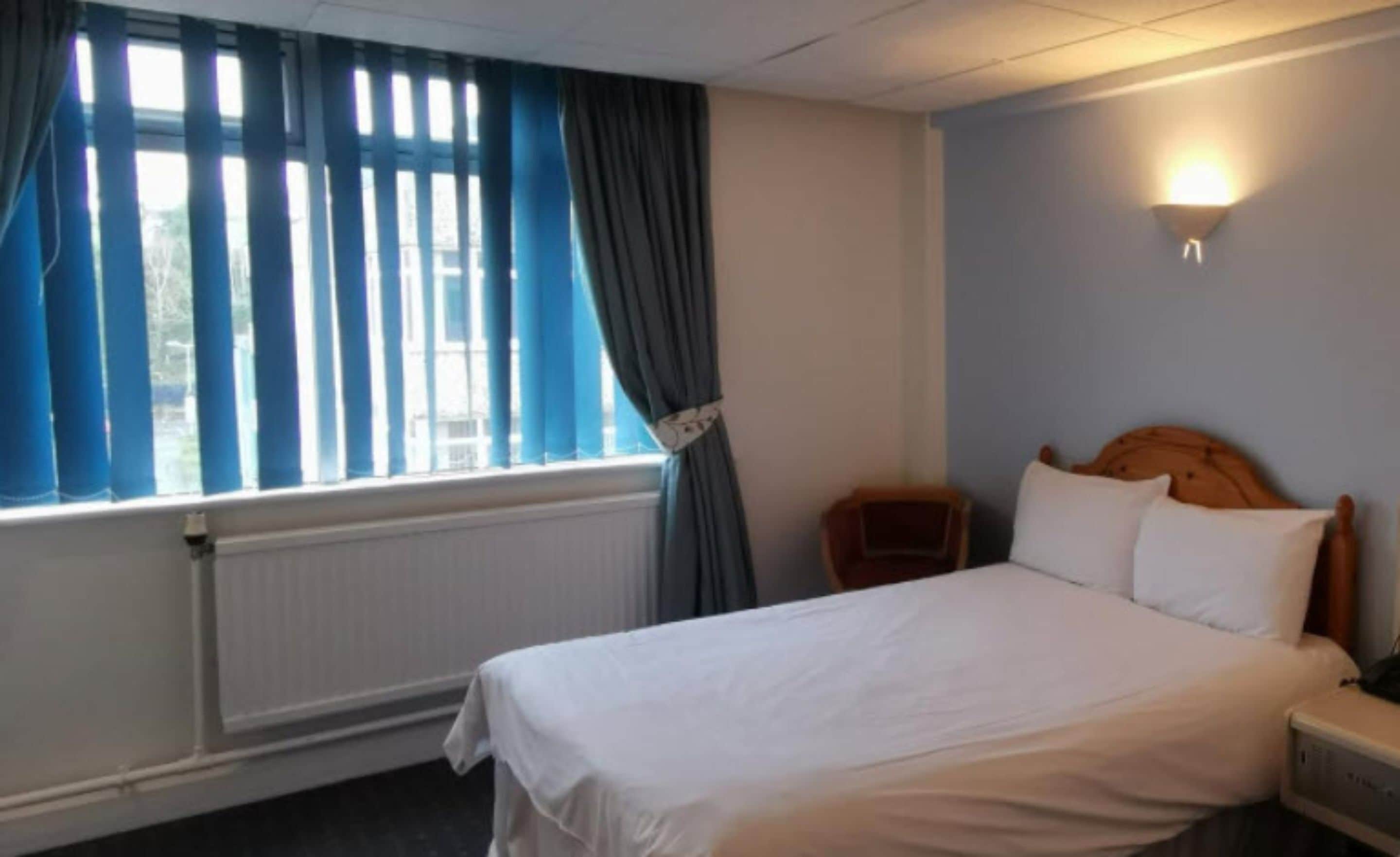 Une chambre d’hôtel avec un lit, une table de chevet, une lampe, une fenêtre avec des rideaux et un radiateur.