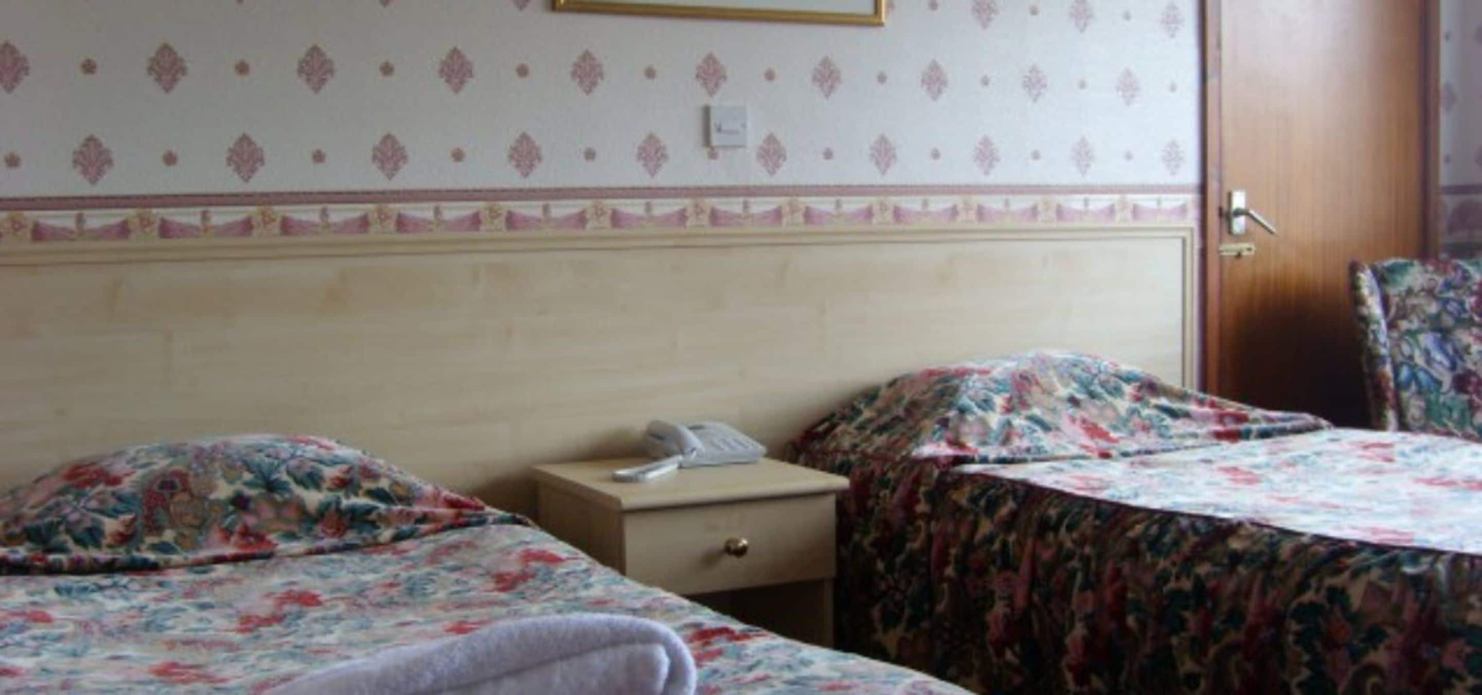 A hotel room with two beds, each with floral bedding, a bedside table with a phone, and a door to the right.