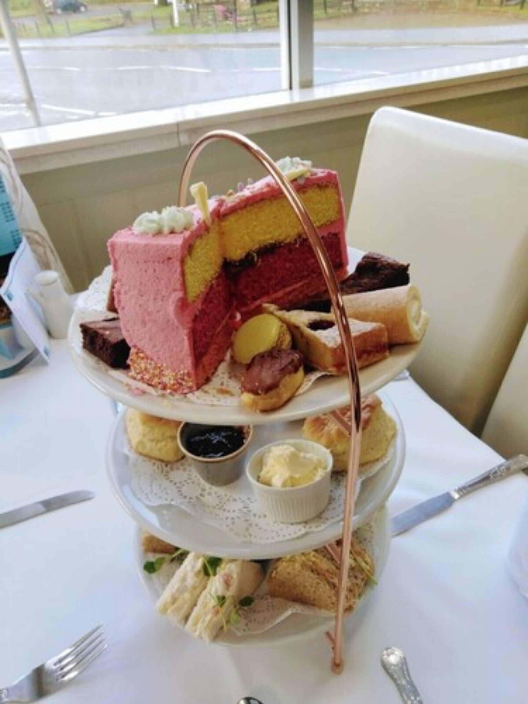 A three-tiered dessert stand with various pastries and a cake, accompanied by small bowls of cream and jam, on a table with a view of the street outside.