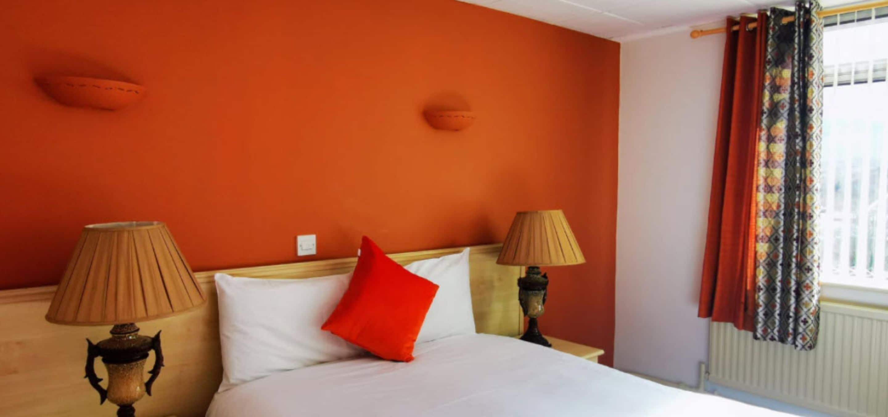 A hotel room with a bed, two bedside lamps, a red pillow, and a window with patterned curtains.