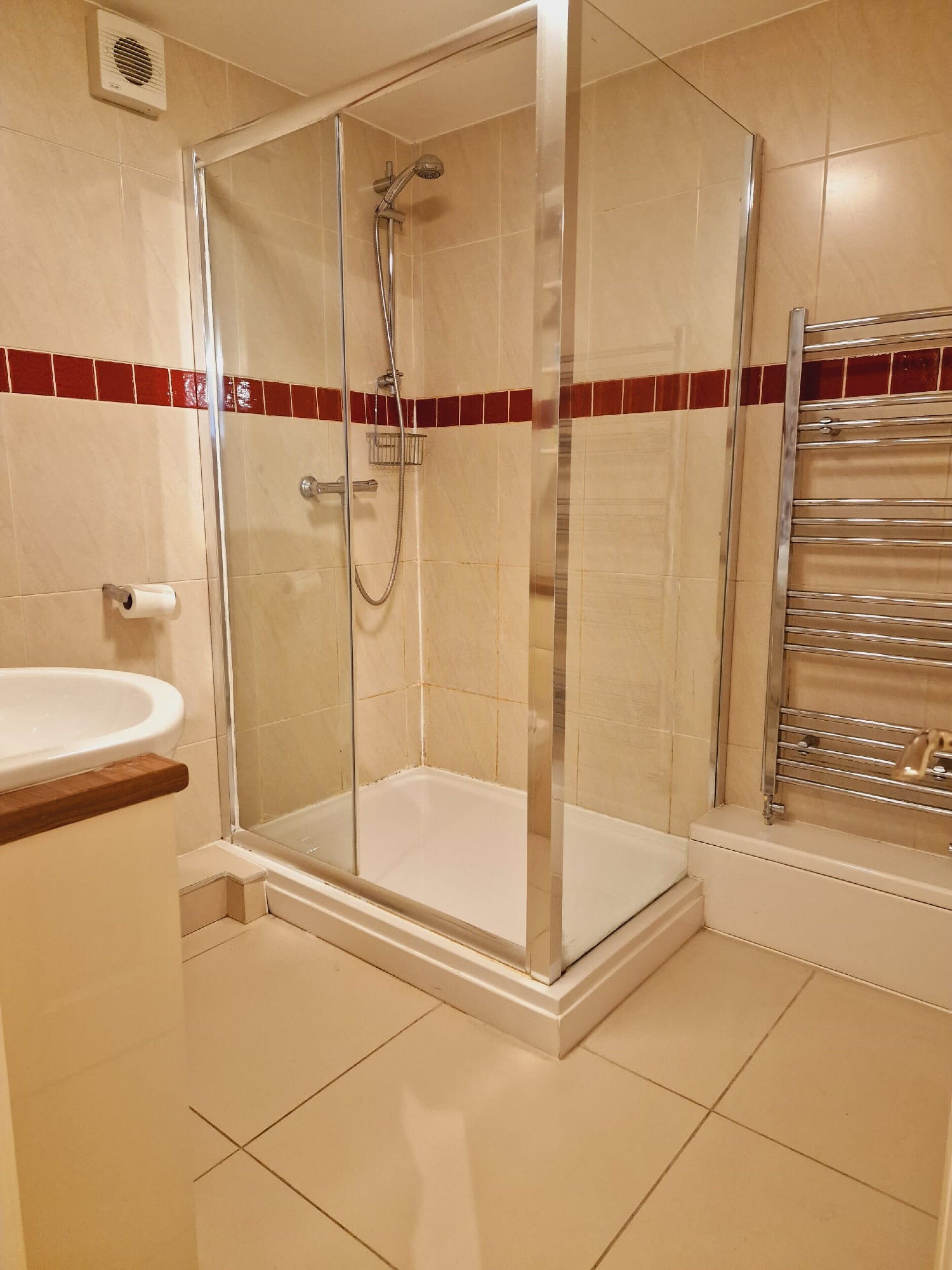 A glass-enclosed shower with a handheld showerhead, a white sink with a wooden countertop, and a towel rack with a towel.