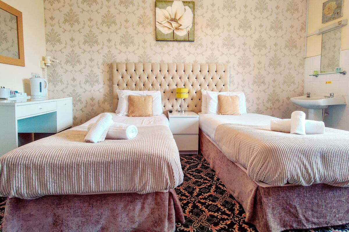Una habitación de hotel con dos camas, un cuadro floral grande en la pared, una mesita de noche con lámpara y un baño con lavamanos y espejo.