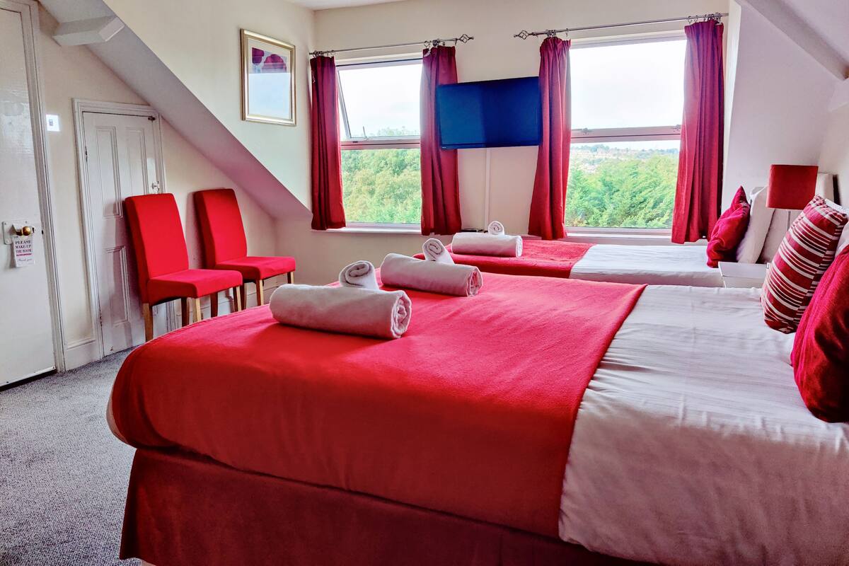 Habitación de hotel con dos camas, ropa de cama roja y blanca, un ventanal con cortinas, un cuadro en la pared y una escalera.
