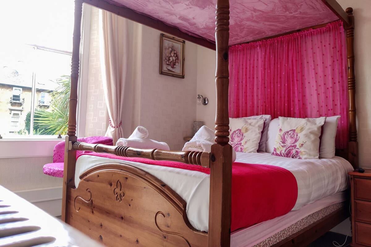 Una cama con dosel de madera y dosel rosa. Hay una ventana con cortinas, un cuadro enmarcado en la pared y una mesita de noche con una lámpara.