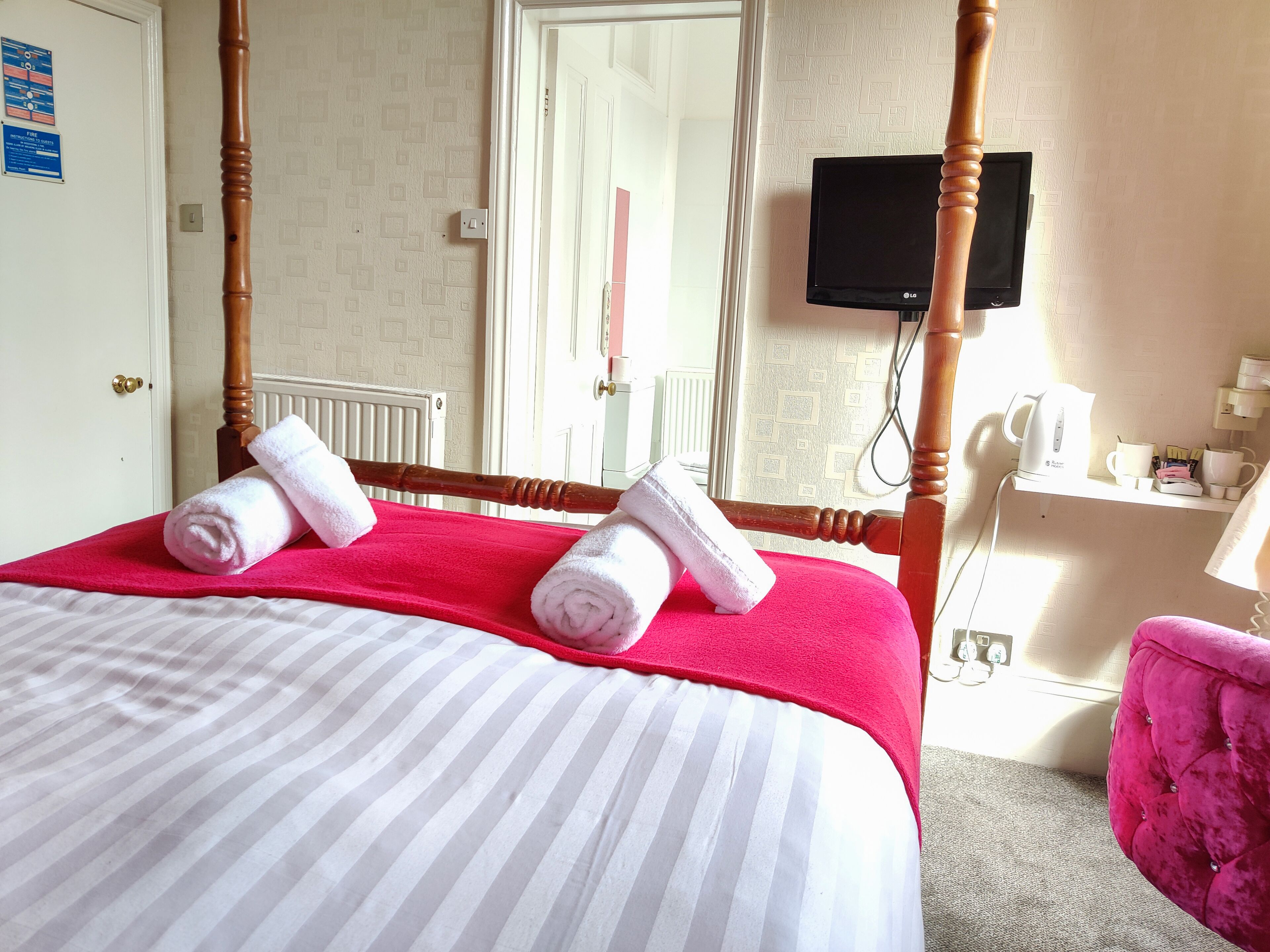 Habitación de hotel con cama con dosel, televisor de pantalla plana, cafetera y sillón rosa.