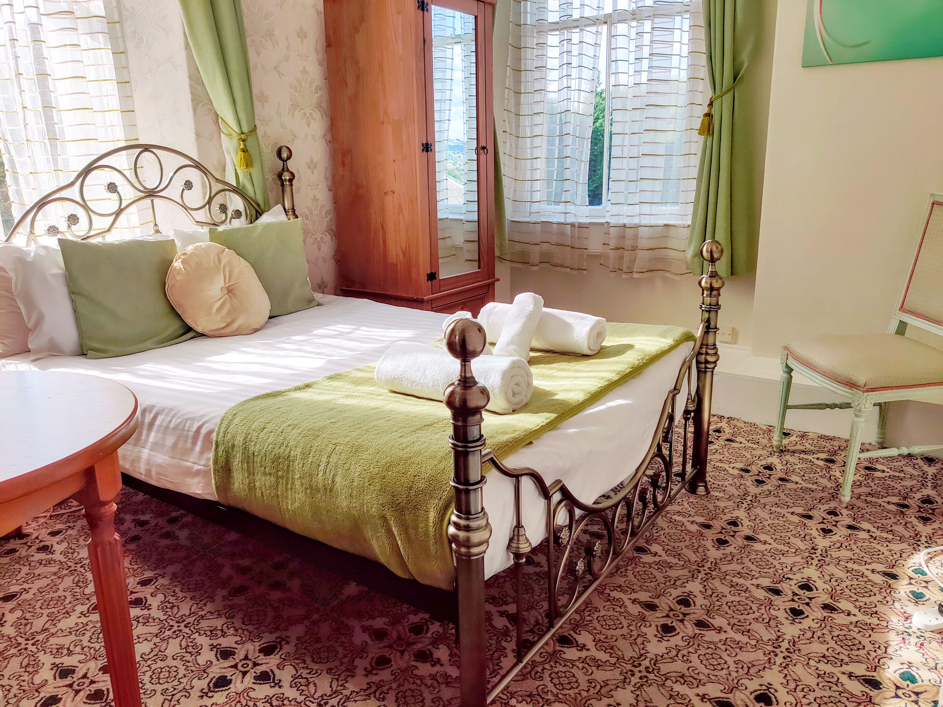 A neatly made bed with pillows and a green blanket, a wooden chair, and a patterned carpet.