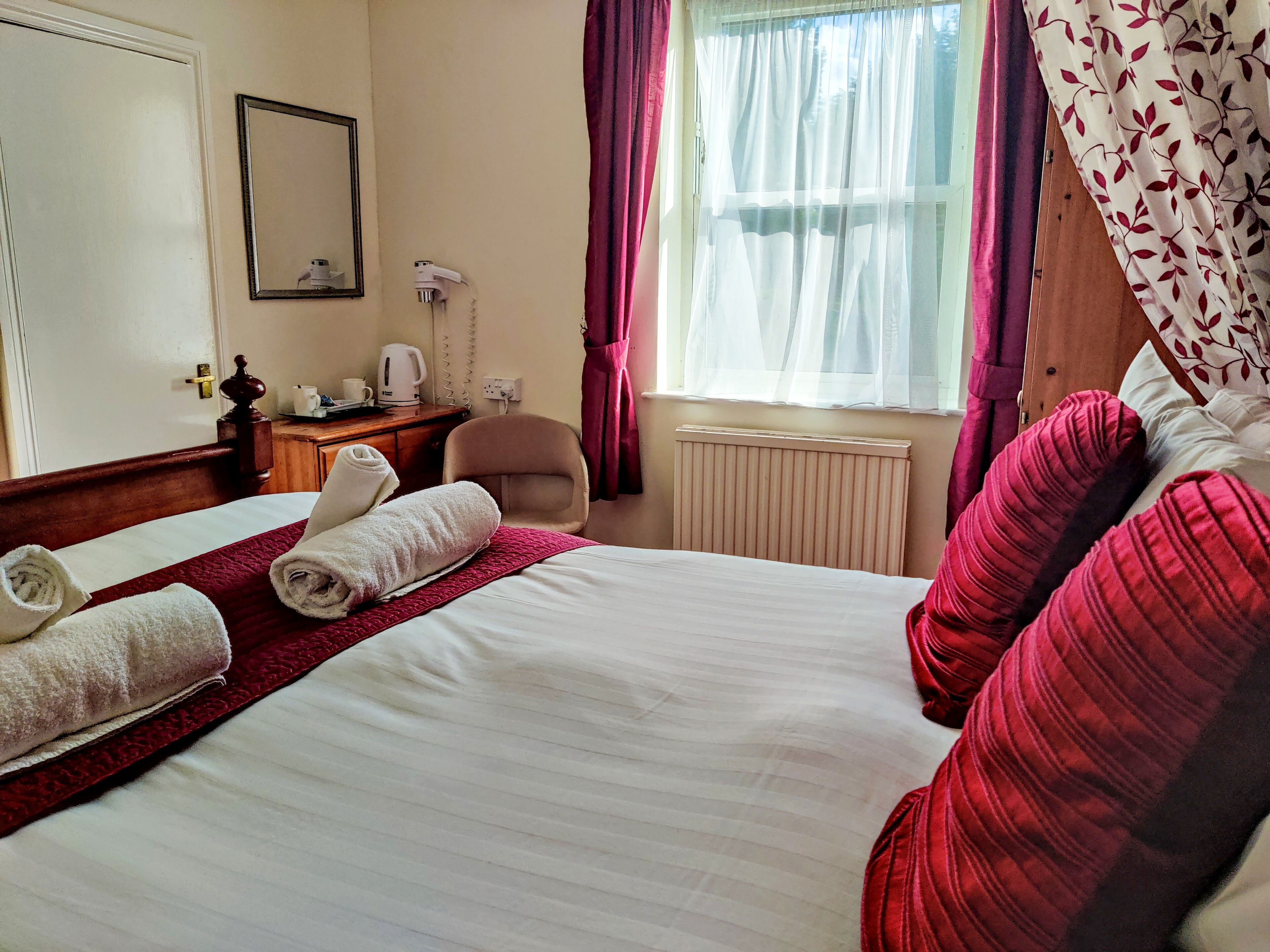A hotel room with a bed, a chair, a desk, a radiator, and a window with curtains.