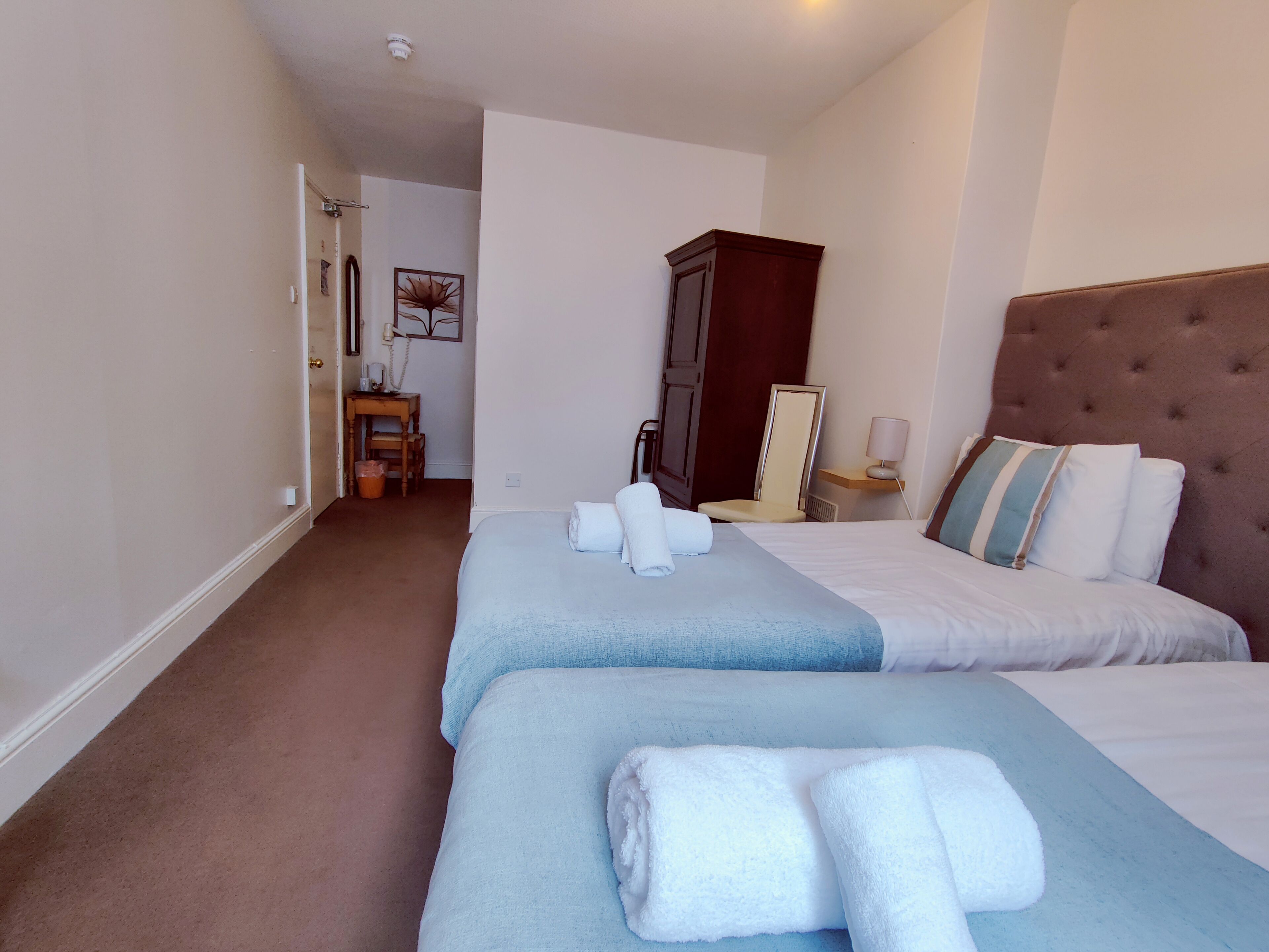 A hotel room with two beds, a wooden wardrobe, a desk with a chair, and a small table with a lamp.