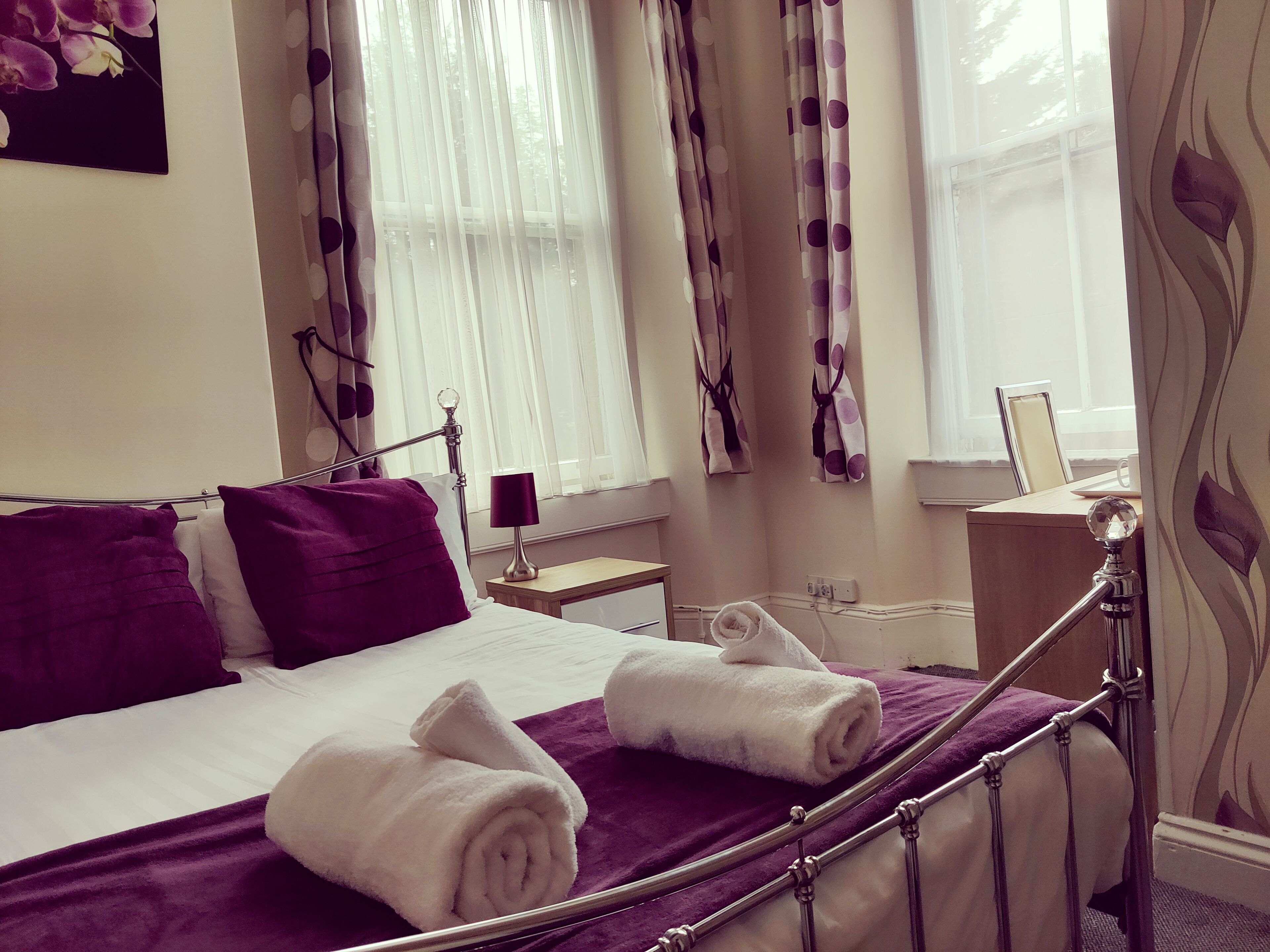 Una habitación de hotel con una cama, almohadas, una mesita de noche, una lámpara y una ventana con cortinas.