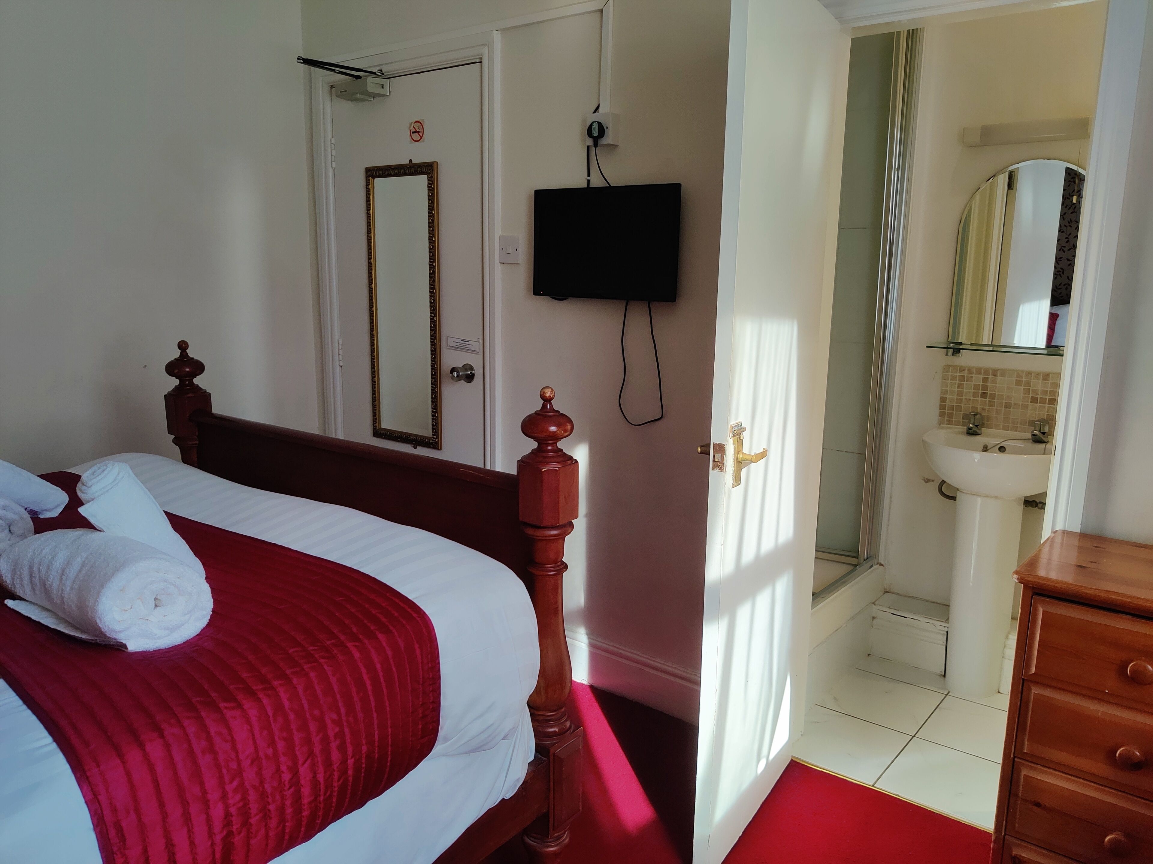 A hotel room with a bed, a TV mounted on the wall, a bathroom with a sink and mirror, and a wooden dresser.