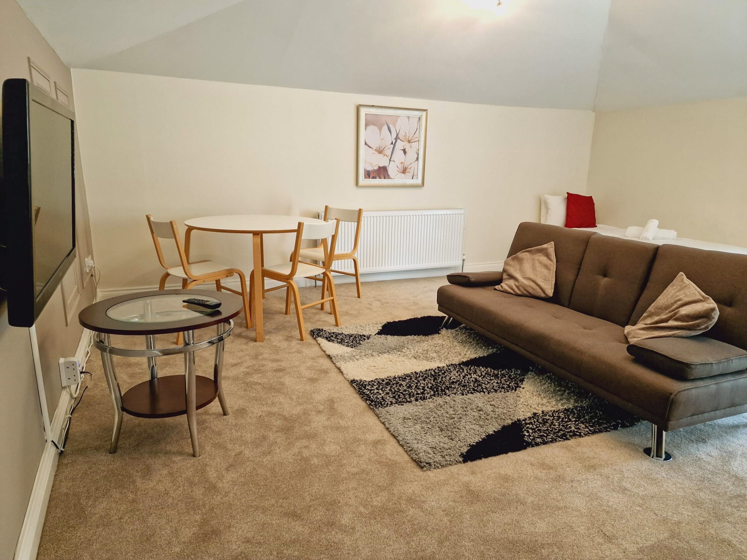 Ein Wohnzimmer mit braunem Sofa, einem Glastisch, Holzstühlen, einem schwarz-weißen Teppich, einem weißen Heizkörper und einem gerahmten Bild an der Wand.