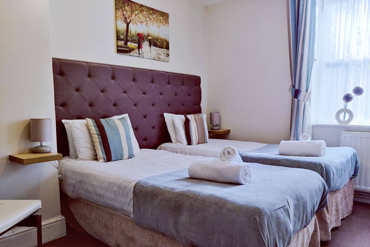 Habitación de hotel con dos camas, un gran cabecero capitonado, lámparas de noche y un cuadro en la pared.