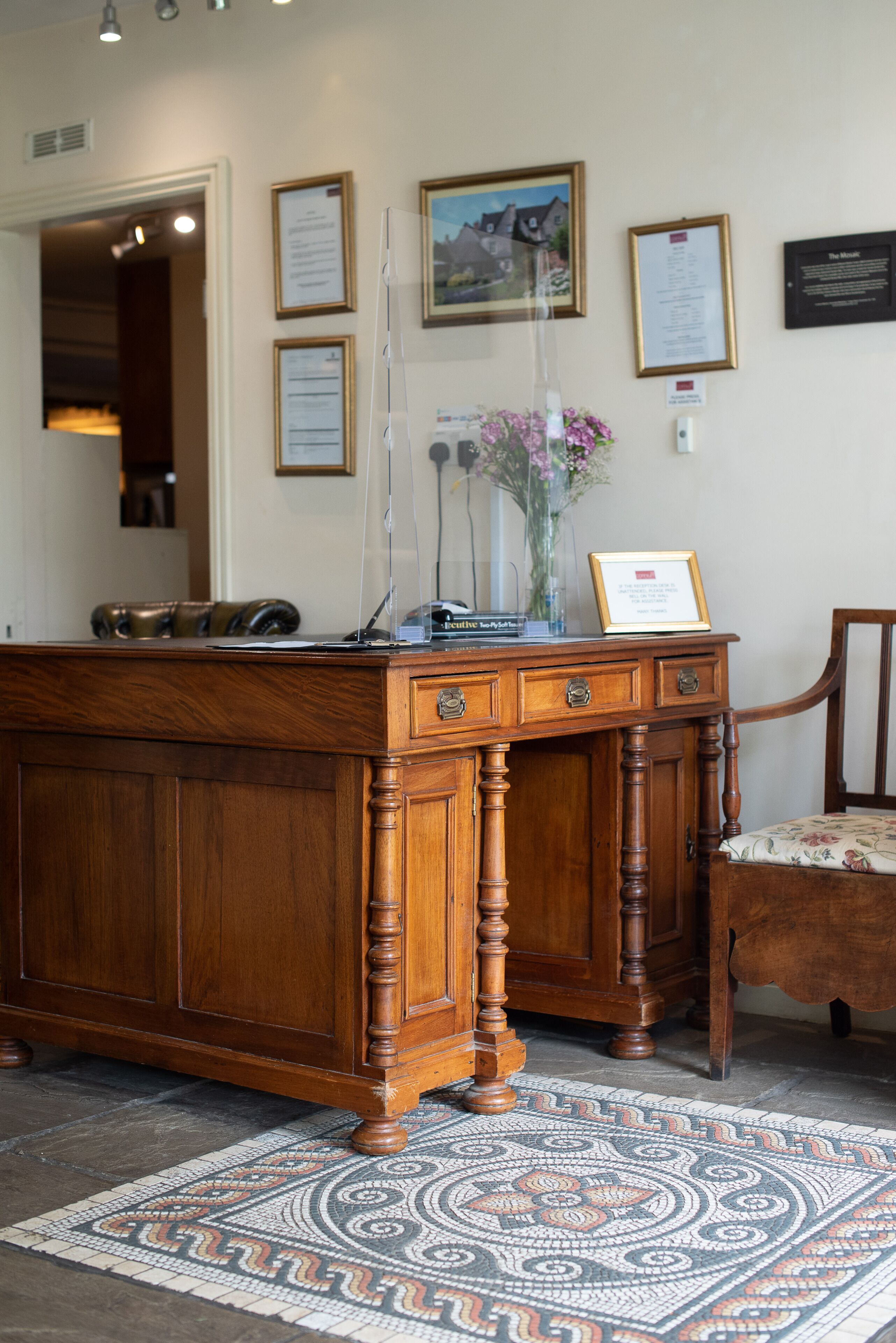 A wooden reception desk with a framed certificate, a chair, and a patterned rug.