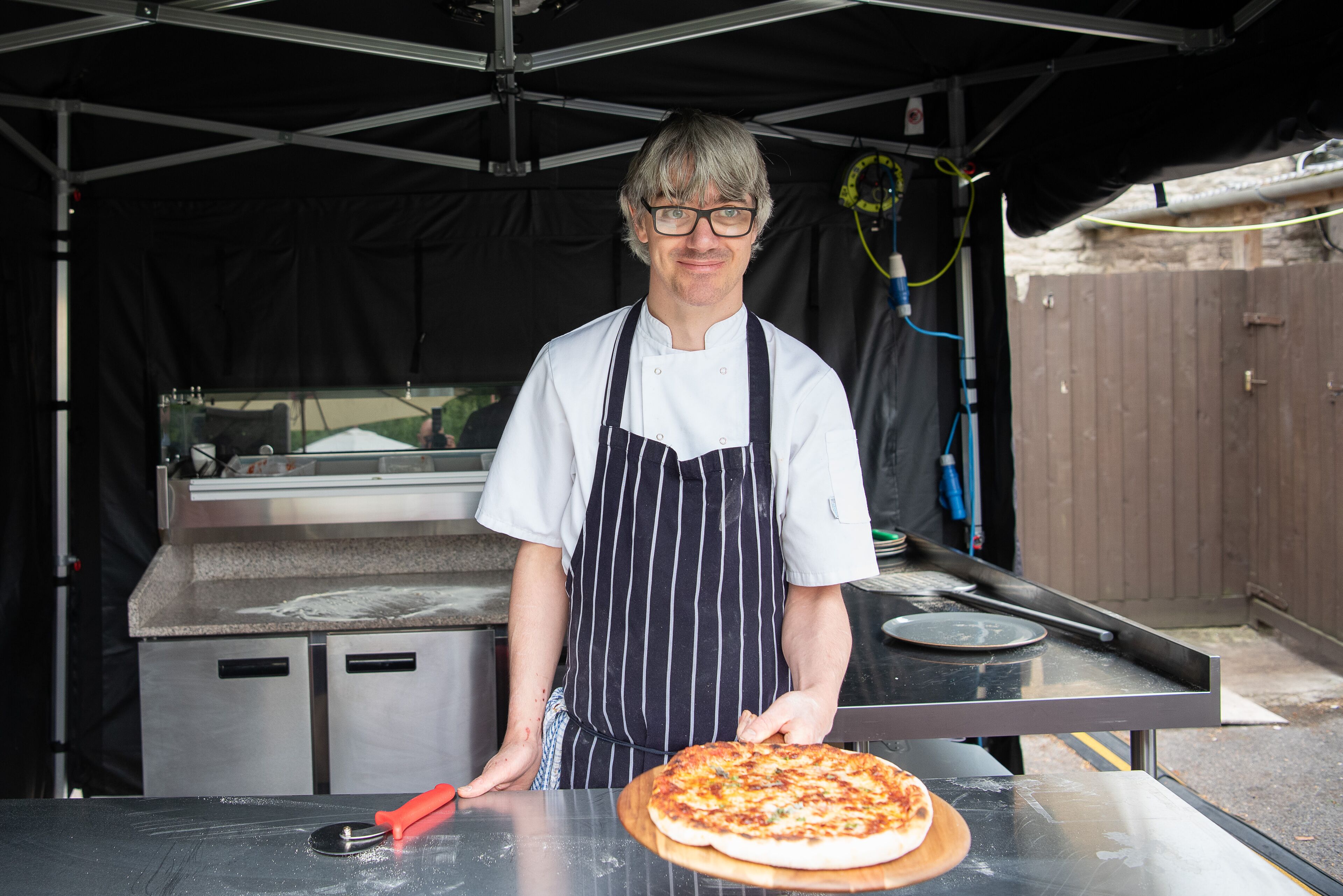 A chef in a white shirt and striped apron holding a pizza on a wooden board.