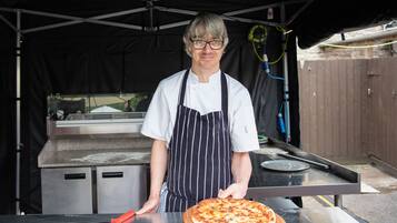 Ein Koch in weißer Kochjacke und gestreifter Schürze hält eine Pizza auf einem Holzbrett.