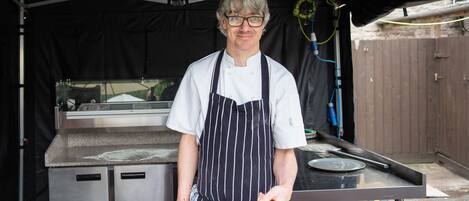 A chef in a white shirt and striped apron holding a pizza on a wooden board.