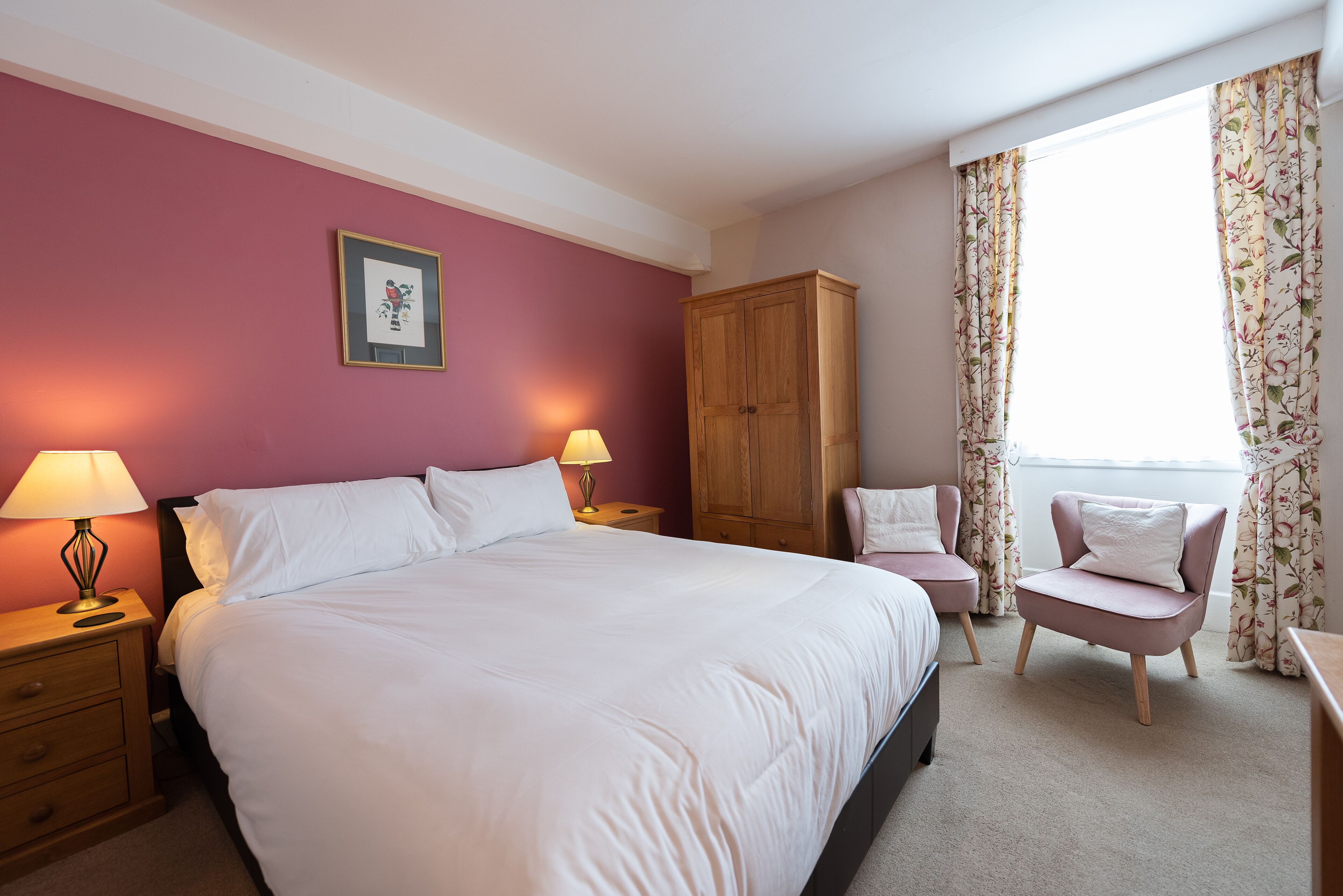 Un dormitorio con una cama grande, dos mesitas de noche con lámparas, un armario de madera, una silla y una ventana con cortinas florales.