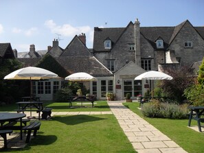 A traditional stone building with a central entrance, outdoor seating area with tables and umbrellas, and a paved pathway. - Corinium Hotel & Restaurant (Cirencester)