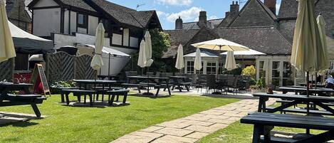 An outdoor seating area with picnic tables and umbrellas, surrounded by buildings with stone chimneys and traditional architecture.