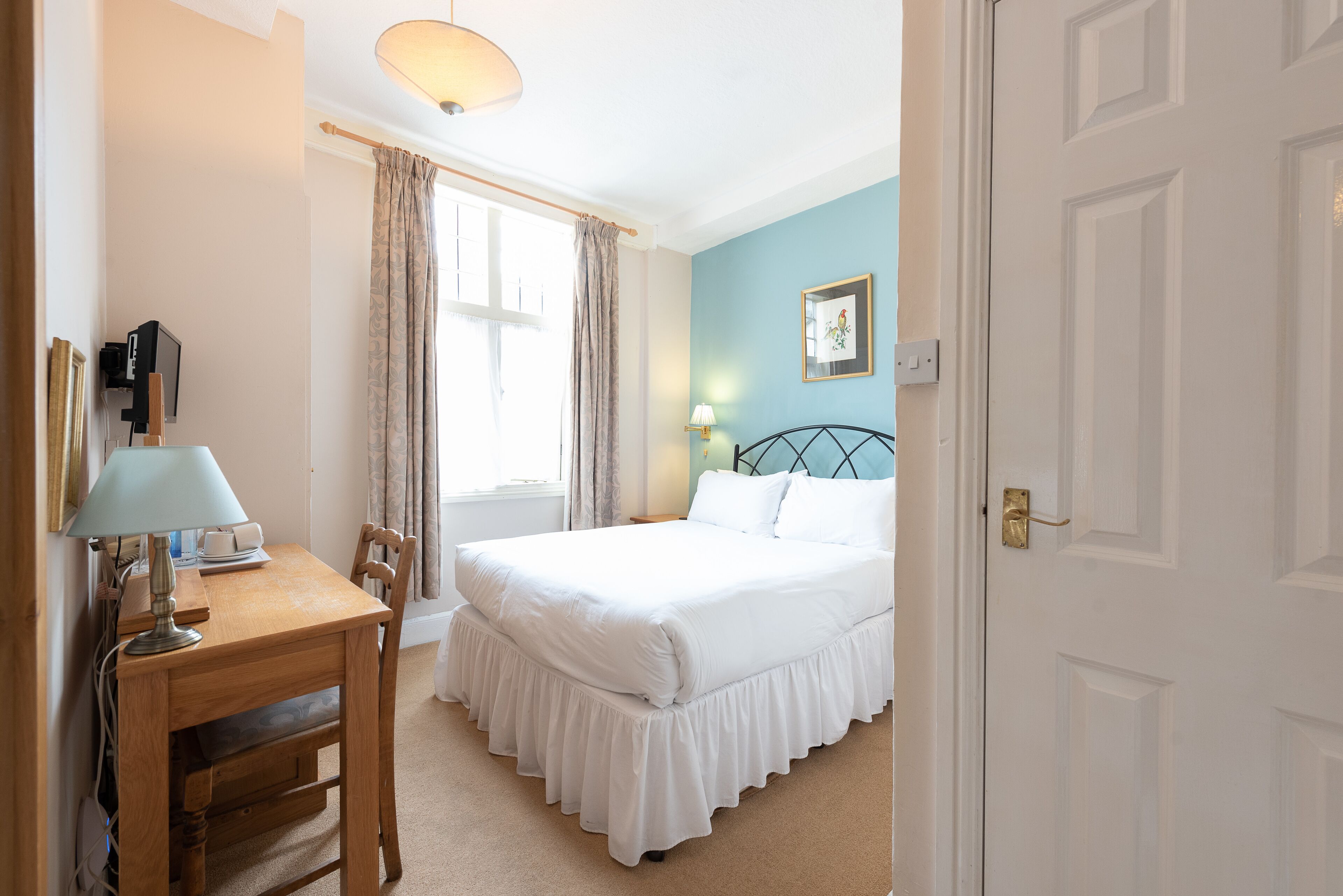A bedroom with a bed, a desk with a lamp, a chair, a window with curtains, and a door.