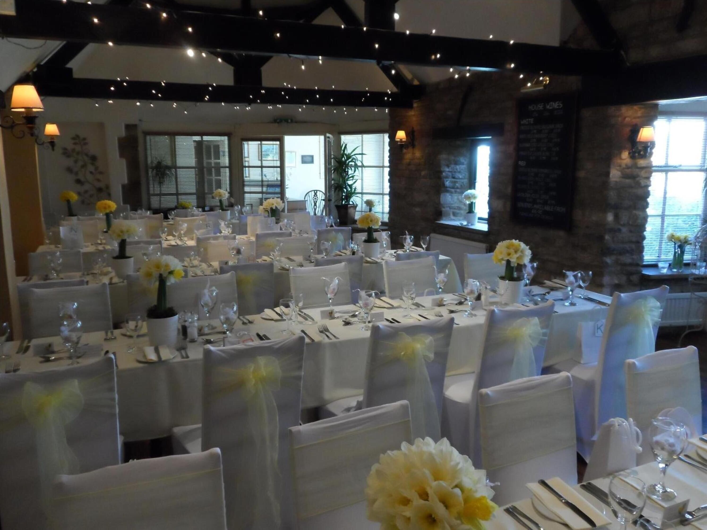A banquet hall with multiple tables set for a formal event, featuring white tablecloths and floral centerpieces.