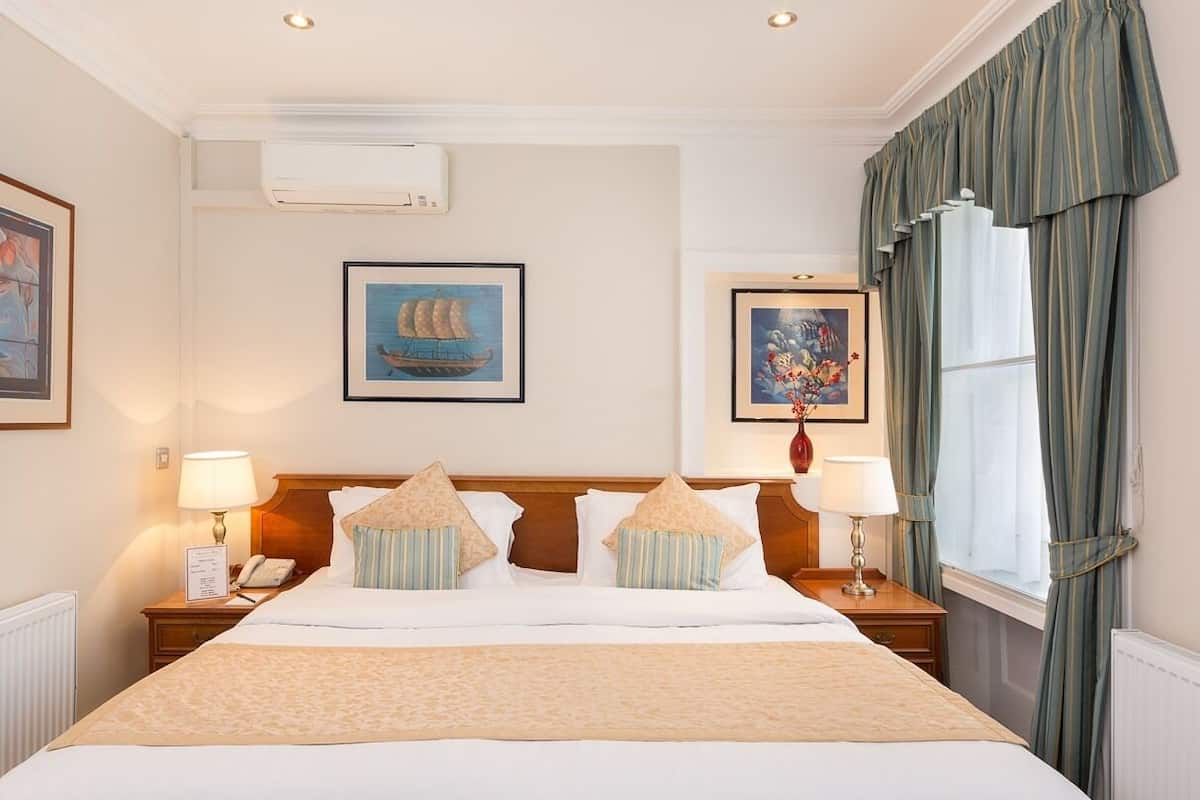 A hotel room with a large bed, two bedside tables with lamps, a window with curtains, and artwork on the walls.