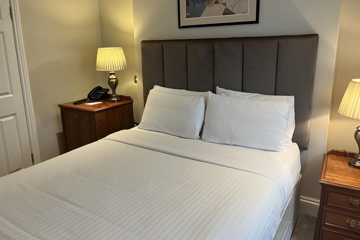 A neatly made bed with white linens, a wooden nightstand with a lamp, and a framed abstract painting on the wall.