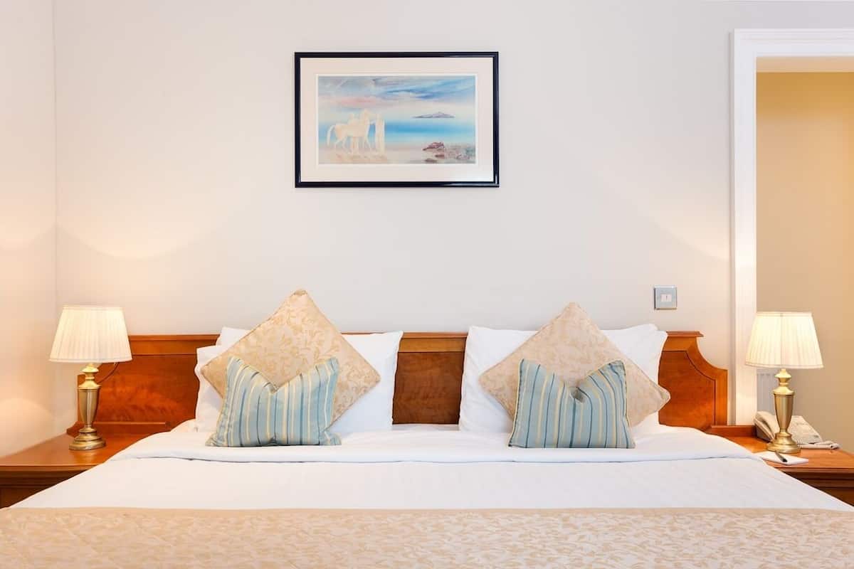 A hotel room with a bed, two bedside lamps, a framed painting on the wall, and a wooden headboard.