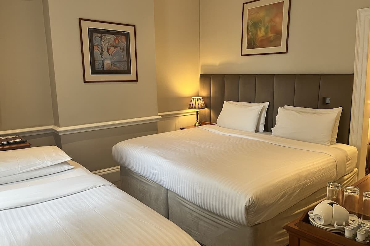 A hotel room with two beds, a headboard, a bedside table with a lamp, and framed artwork on the walls.