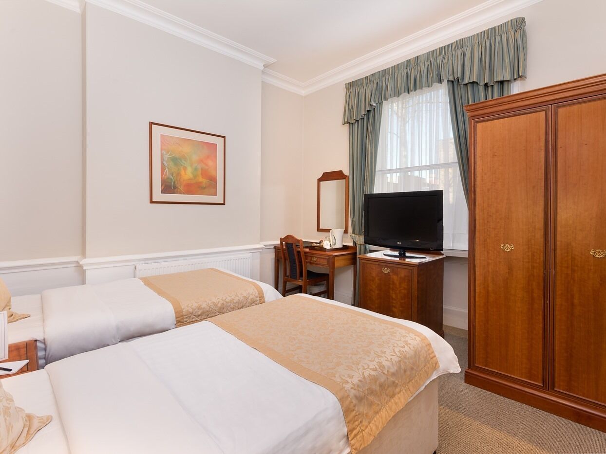 A hotel room with two beds, a television, a desk with a chair, a wardrobe, and a painting on the wall.