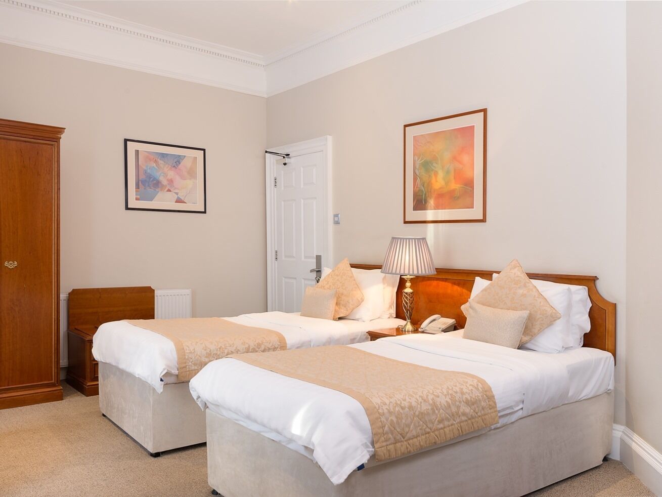A hotel room with two beds, a wardrobe, a radiator, and two framed artworks on the walls.