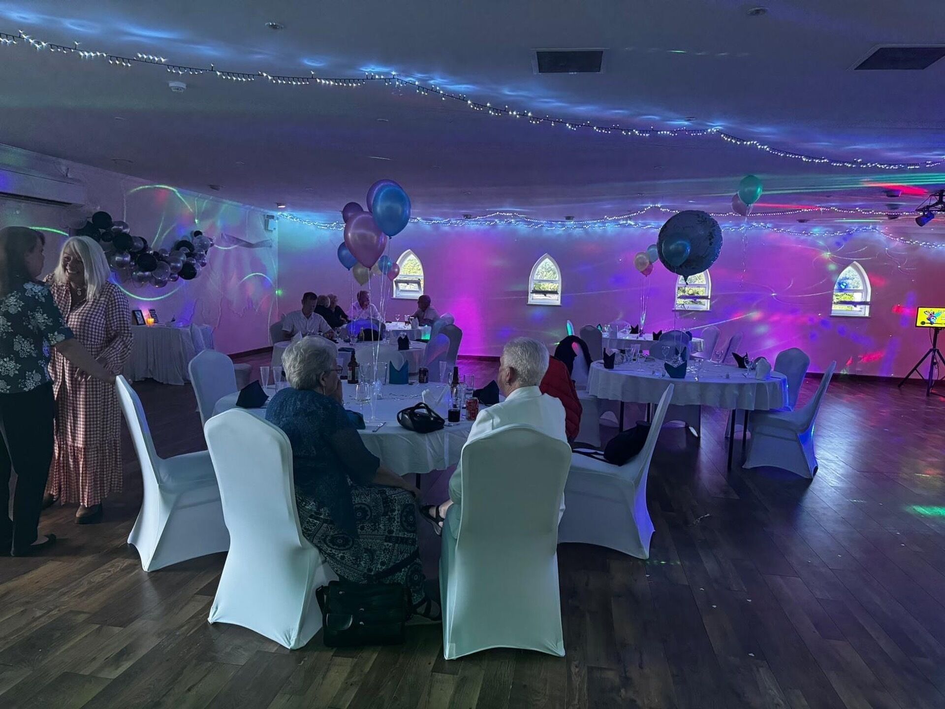 Une salle avec des tables et des chaises rondes, décorée de ballons et de guirlandes lumineuses.