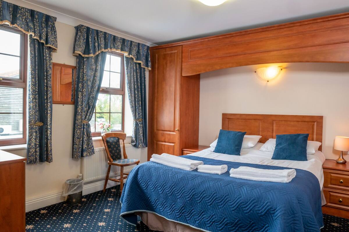 A hotel room with a large bed, wooden headboard, and a nightstand with a lamp. There is a window with curtains and a chair near the window.