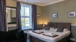 A neatly made bed with pillows and a folded towel, a bedside table with a lamp, a chair, and a window with curtains. - Shurland Hotel (Sheerness)