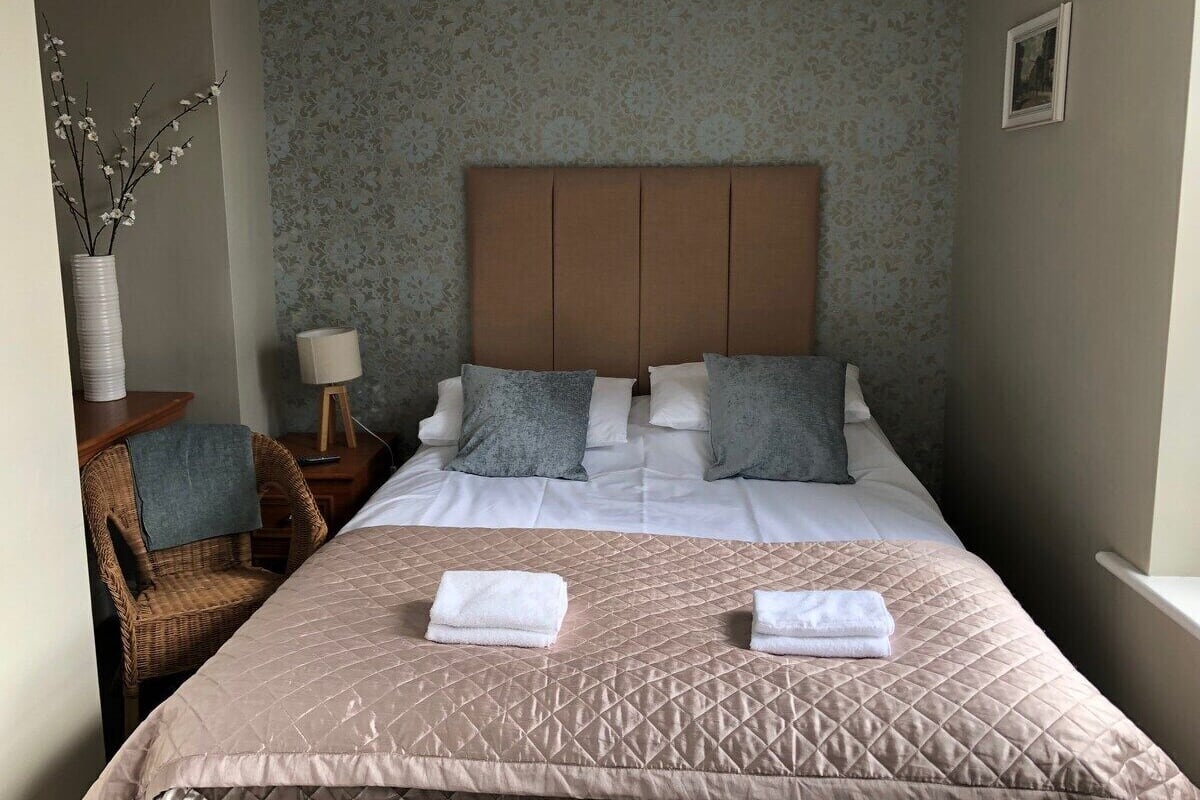 A neatly arranged hotel room with a bed, pillows, a chair, a lamp, and a vase with flowers.