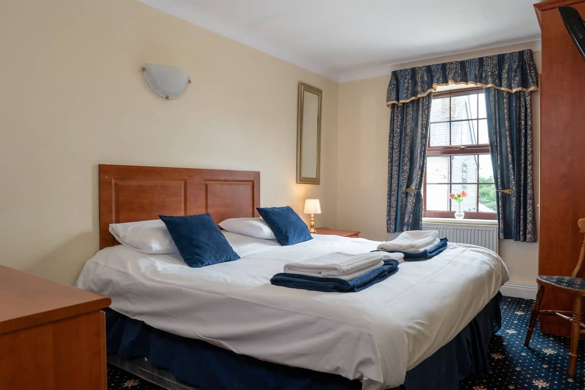 A hotel room with a wooden headboard, two beds with white and blue bedding, a nightstand with a lamp, a window with curtains, and a wooden chair.