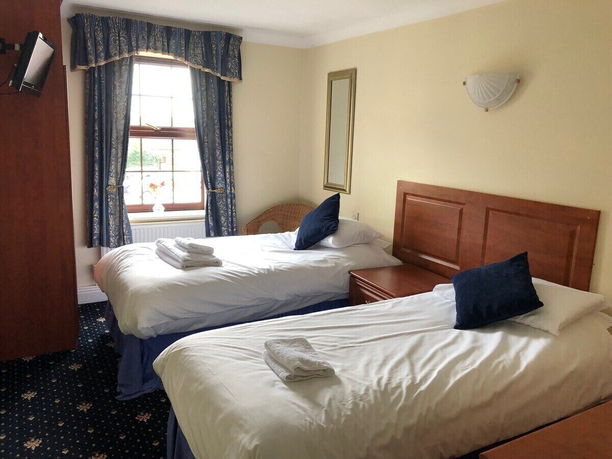 A hotel room with two beds, a window with curtains, a mirror, and a wall-mounted light fixture.