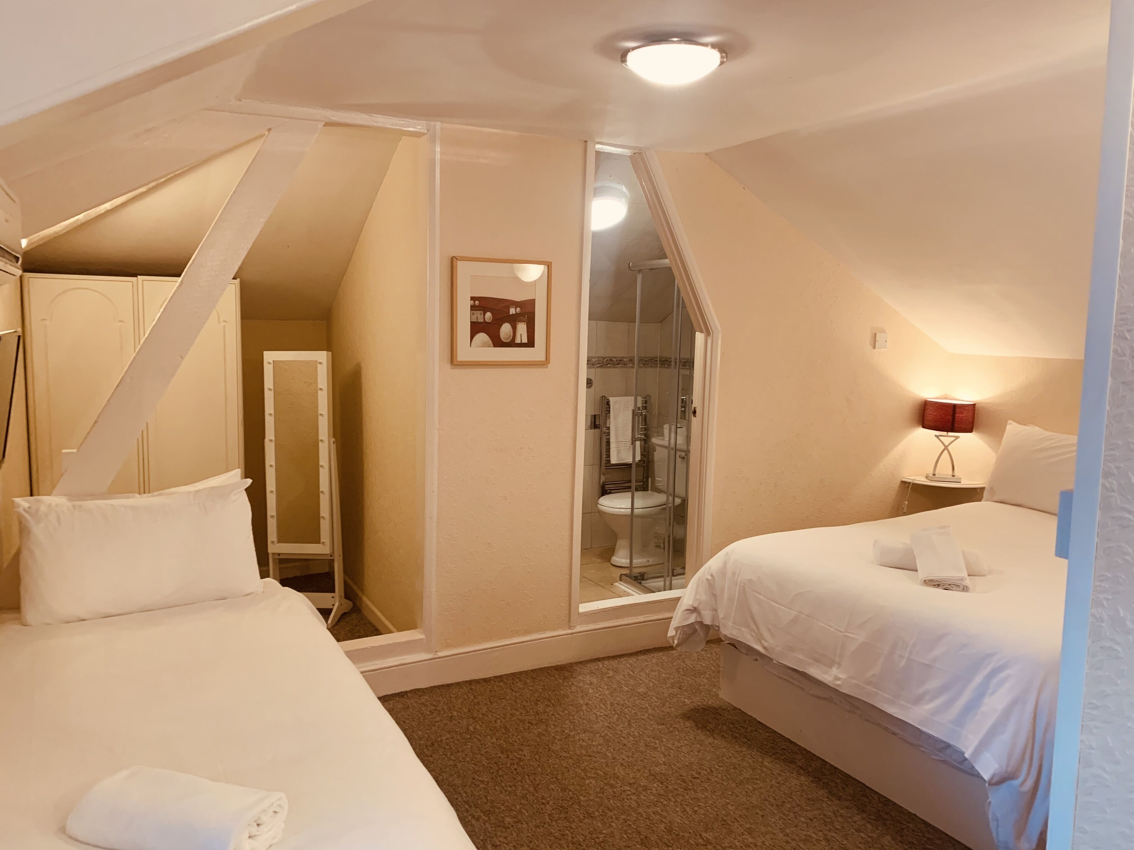 Quarto de hotel com duas camas, banheiro visível através de uma porta de vidro e uma pequena mesa de cabeceira com abajur.