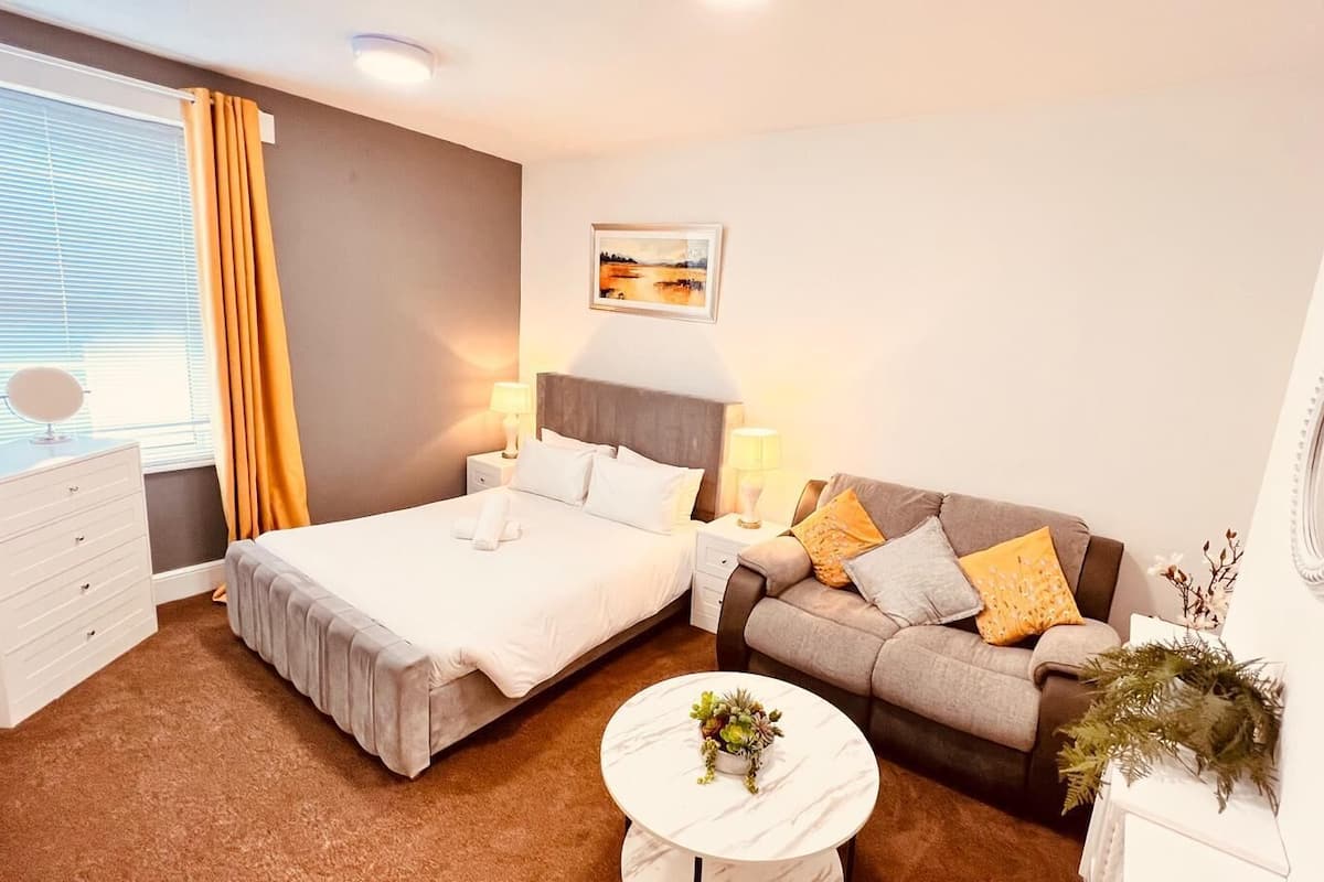 A hotel room with two beds, a sofa, a bedside table, a lamp, a mirror, and a window with curtains.