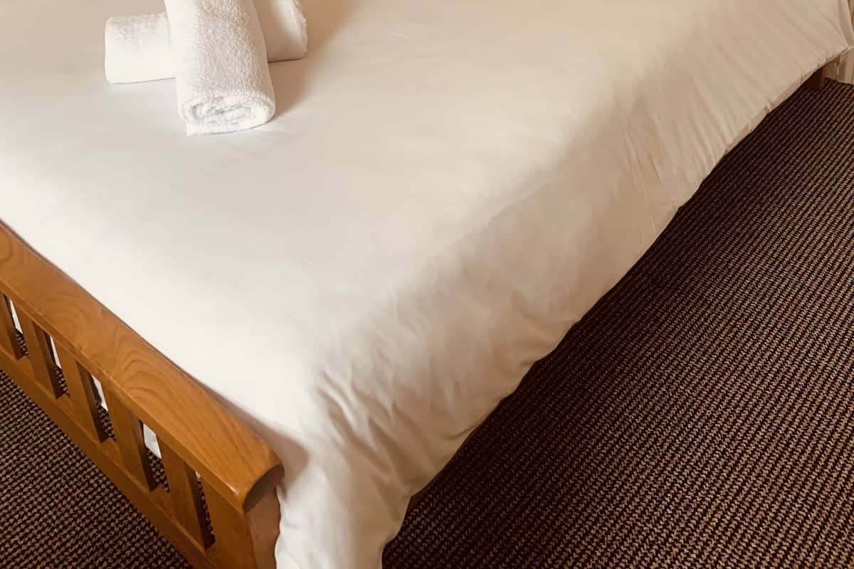 A neatly made bed with white linens and a rolled towel, a wooden headboard, and a brown carpeted floor.