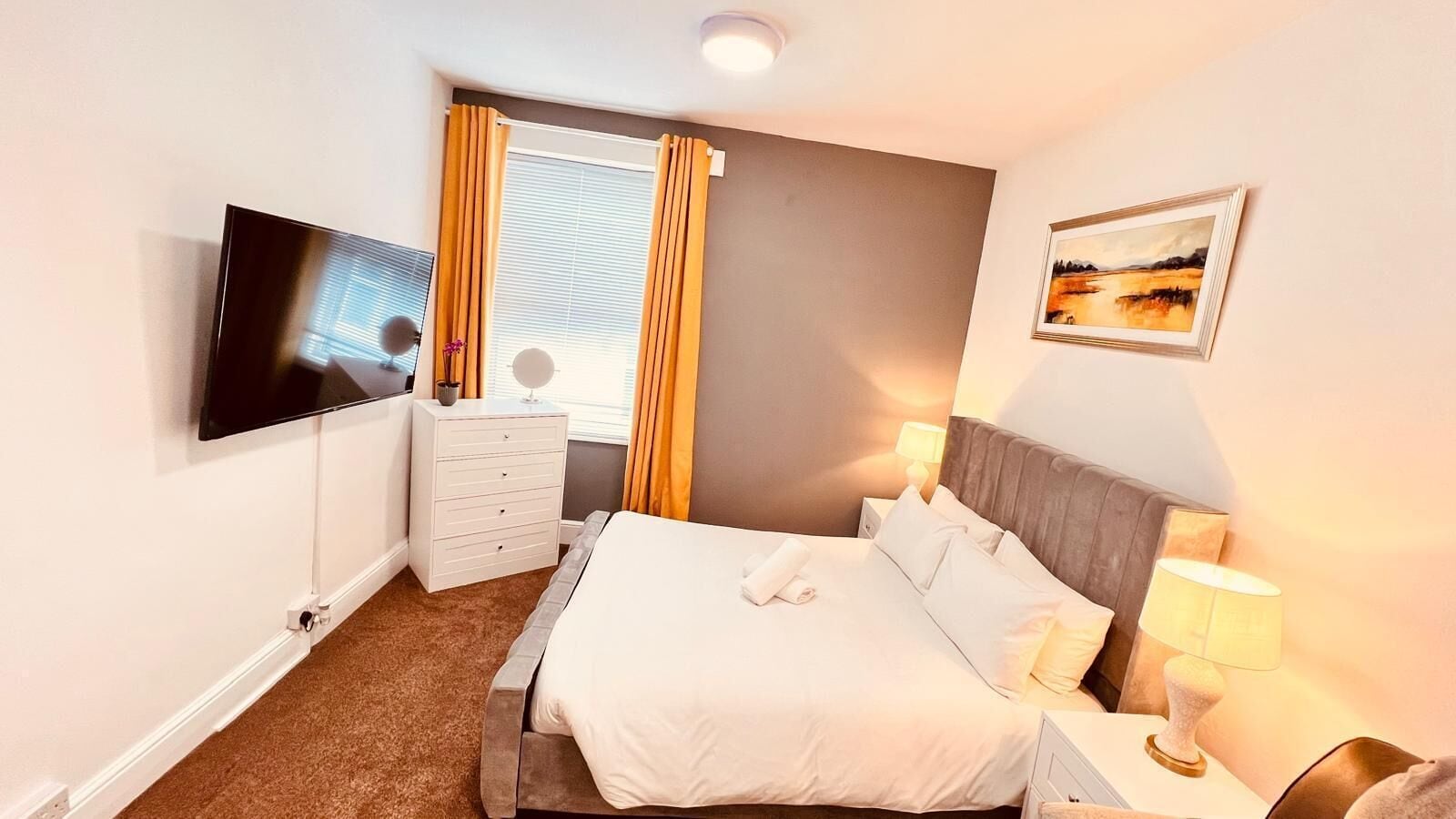 A hotel room with a bed, a TV, a dresser, and a painting on the wall.