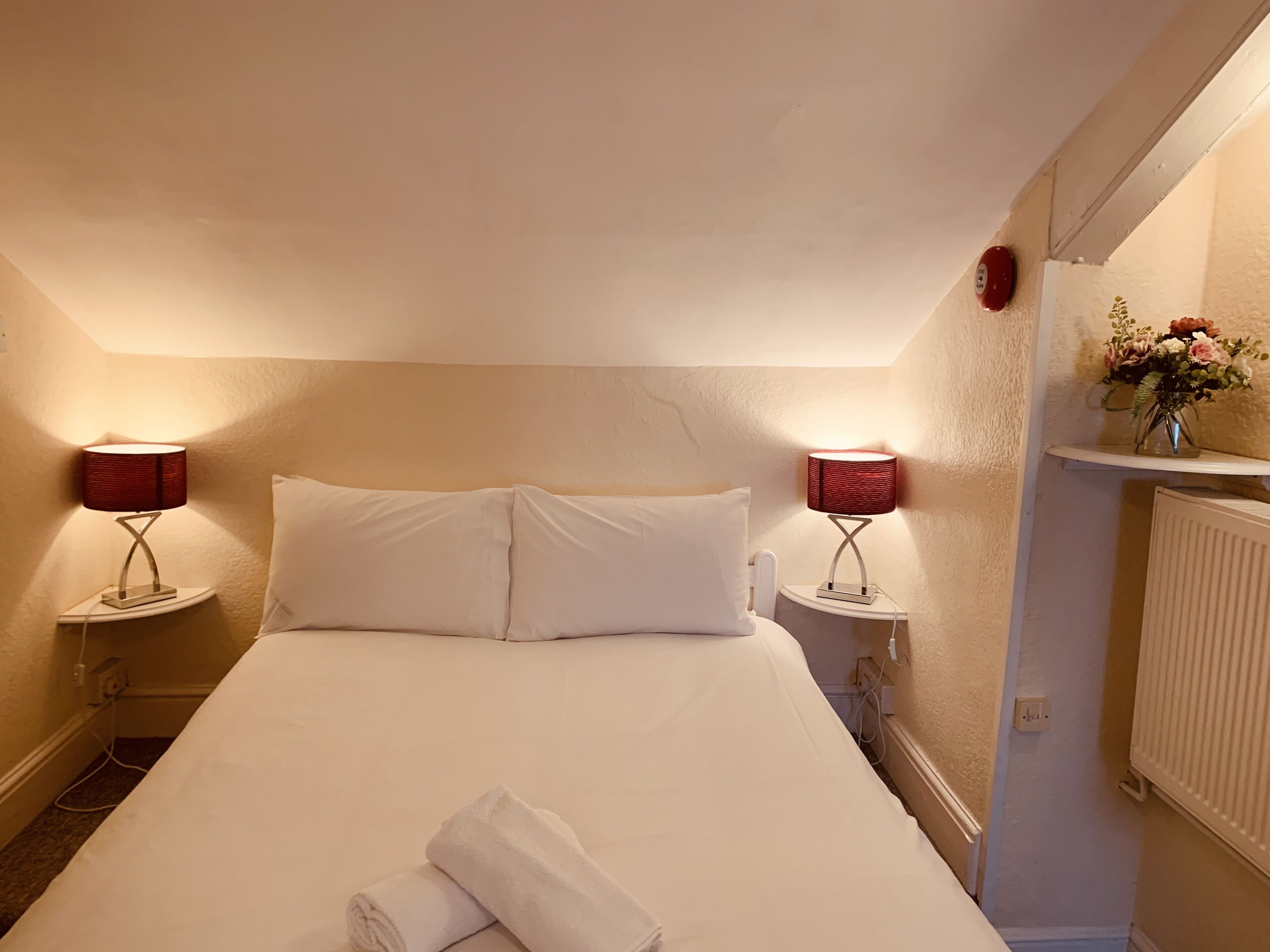A neatly made bed with white linens, two bedside lamps, a small vase with flowers, and a radiator on the wall.