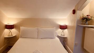 A neatly made bed with white linens, two bedside lamps, a small vase with flowers, and a radiator on the wall.