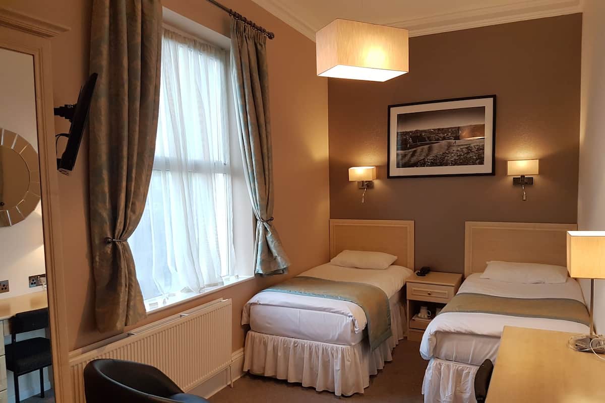 Ein Hotelzimmer mit zwei Betten, einem Sessel, einem Schreibtisch, einem Heizkörper und einem Fenster mit Vorhängen.