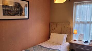 A hotel room with a bed, a nightstand with a lamp, a framed picture on the wall, and a window with curtains.