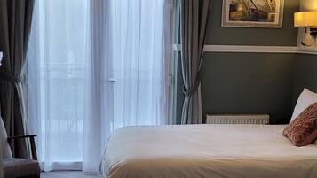 Quarto de hotel com uma cama grande, uma cadeira, uma escrivaninha e uma janela com cortinas.
