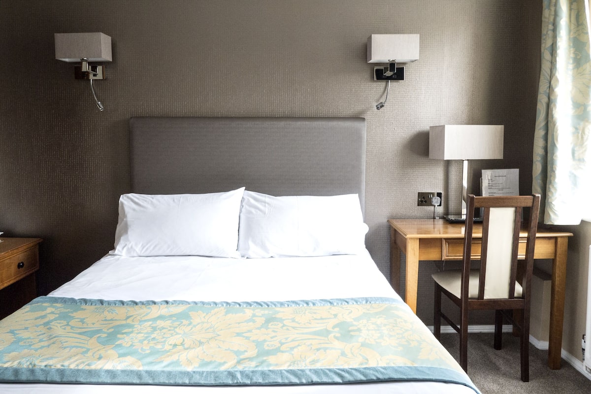 Ein Hotelzimmer mit einem großen Bett, zwei Nachttischlampen, einem Holznachttisch, einem Schreibtisch mit Stuhl und einem Fenster mit Vorhängen.
